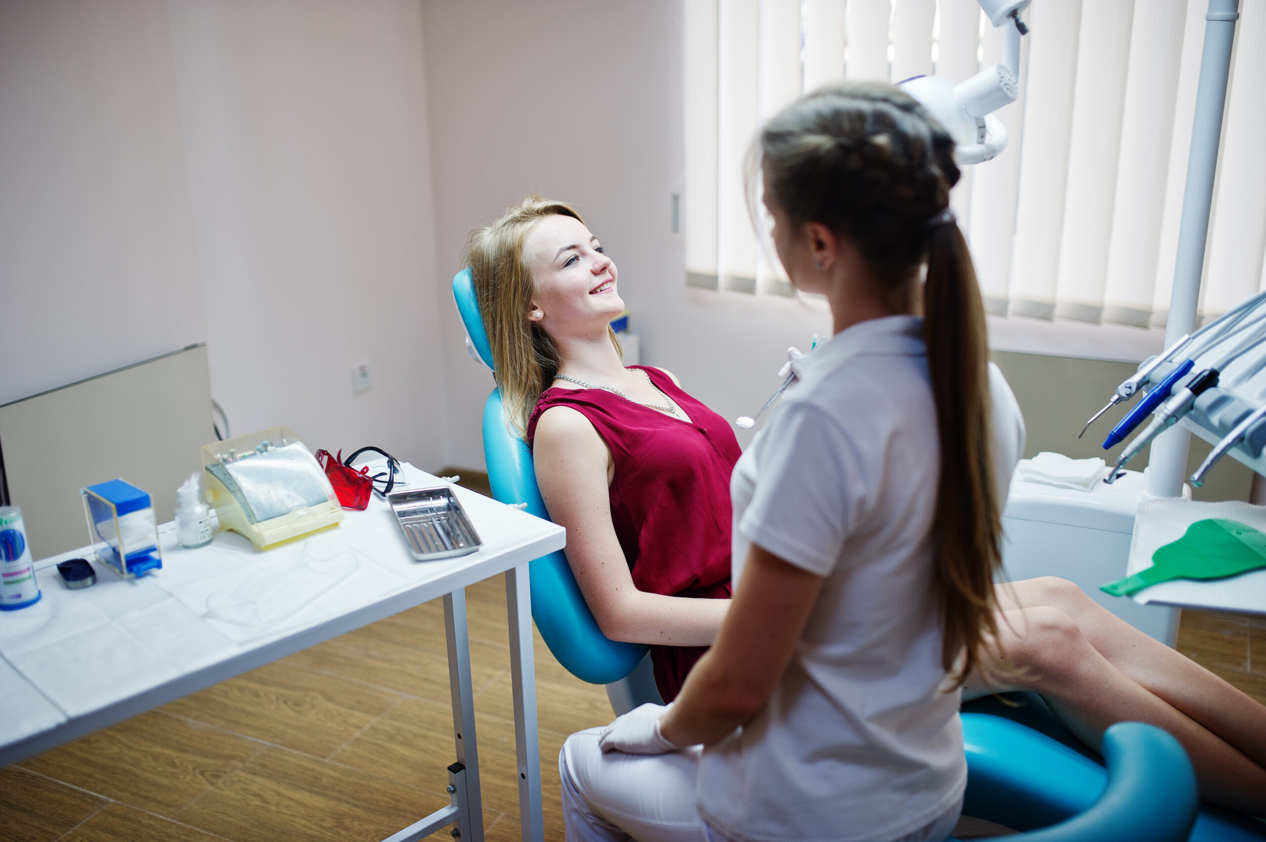 Attractive patient in red-violet dress laying on the dental chair while female dentist treating her teeth with special instruments.
